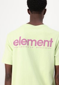 Světl zelené tričko s krátkým rukávem, které má na zádech velké fialové logo "element" a text "SKATEBOARD CO. WORLDWIDE". Měkká textilie.