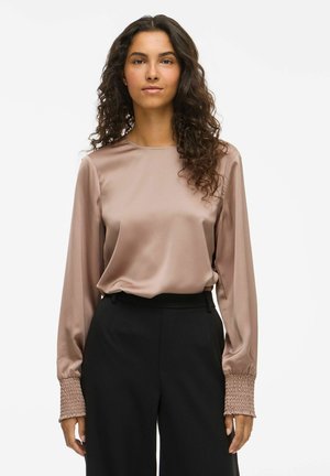Woman with long curly hair wearing a shiny mauve blouse with textured cuffs and high-waisted black trousers, standing against a white background.