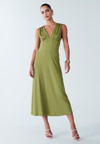 Groene, mouwloze midi-jurk met een diepe halslijn, geplooide lijfje en een getailleerde taille. Zachte stof met een subtiele textuur, gecombineerd met witte hakken.