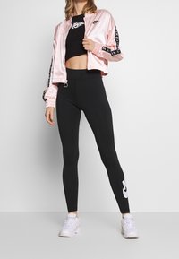 Roze satijnen bomberjack met zwarte accenten, cropped design, gecombineerd met hoge zwarte leggings en witte sneakers.