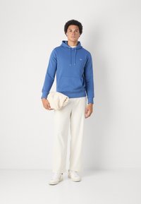 Jonge man met een blauwe hoodie, witte broek en witte sneakers, staand en een gevouwen beige jas vasthoudend tegen een effen achtergrond.