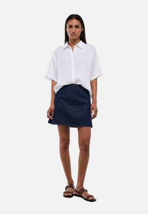 Femme debout portant une chemise blanche ample à manches courtes avec des boutons, une jupe en jean bleu foncé et des sandales marron foncé sur un fond uni.