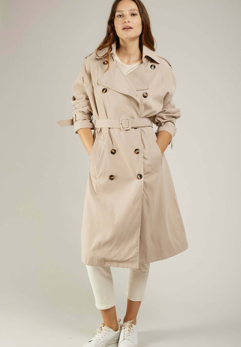 Manteau beige à double boutonnage, avec ceinture et manches retroussées. Tissu lisse, design midi, et poches latérales.