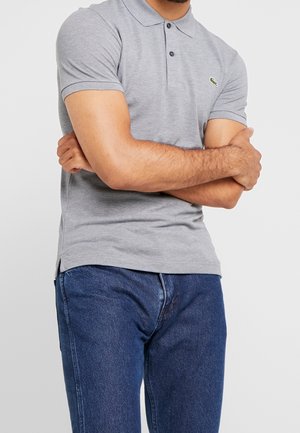 Mężczyzna w szarej koszulce polo z krótkim rękawem i logo, w niebieskich dżinsach, z założonymi na piersiach rękami, na jednolitym jasnym tle.