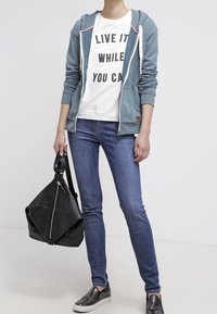 Felpa blu con zip sopra una t-shirt bianca con testo nero. Indossata con jeans blu aderenti, borsa in pelle nera e scarpe slip-on con motivo.