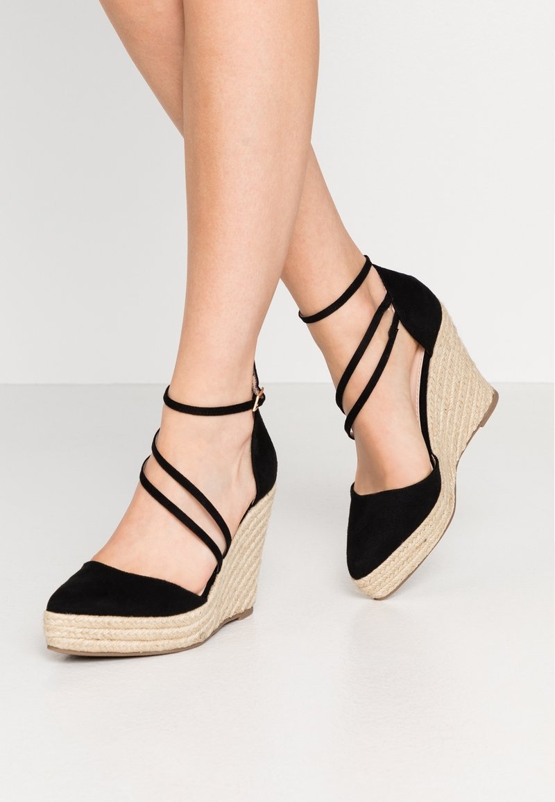 Jambes portant des espadrilles compensées noires avec des brides à la cheville et des talons compensés enveloppés de jute sur un fond gris clair.