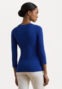 Blue long-sleeve top with a round neckline, smooth texture, and fitted silhouette, paired with light-colored trousers.