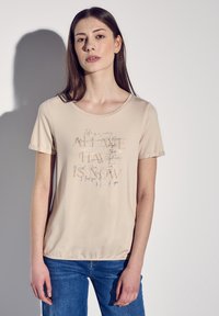 T-shirt beige à manches courtes avec un col rond, présentant un motif texturé de citations dans des polices variées et des accents argentés.