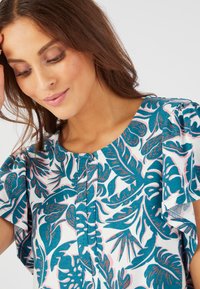 Blusa de manga corta con un estampado floral, que presenta hojas en tonos verde azulado y rosa sobre un fondo blanco, con escote redondo y detalles de botones.