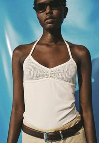 Osoba nosící tmavé sluneční brýle, bílý halter top s perforovanými detaily nahoře, béžové kalhoty a hnědý opasek na modrém pozadí.