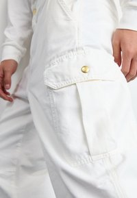 Salopette en coton blanc avec une finition texturée, dotée d'une poche cargo latérale fermée par un bouton pression doré et de manches longues.