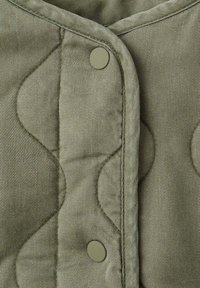 Olivově zelená prošívaná látka s texturovaným vzorem, obsahující řadu strojově šitých křivek a kulaté patentové zapínání.