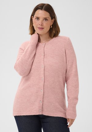 Frau mit schulterlangen braunen Haaren, die goldene Creolen und eine rosa Strickjacke mit Knöpfen trägt, posiert mit der Hand am Hals vor weißem Hintergrund.
