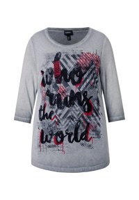 T-shirt gris à manches longues arborant un design graphique avec le texte « who runs the world » en noir et rouge, sur un fond texturé avec des motifs.