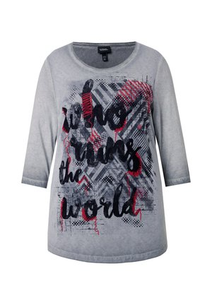 Harmaa pitkähihaisen t-paita, jossa on graafinen suunnittelu tekstillä "kuka hallitsee maailmaa" mustalla ja punaisella, tekstuurilla taustalla, jossa on kuvioita.