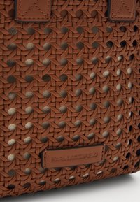 Brahno tkana torba iz teksturiranega materiala, ki ima gladko usnjeno oznako z vtisnjenim napisom "KARL LAGERFELD" na dnu.