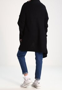 Cardigan noir en tricot surdimensionné avec une texture côtelée, présentant une coupe ample. Associé à un jean bleu à revers et des baskets blanches.