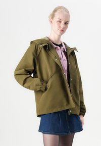 Olive green hooded jacket with snap buttons, elastic cuffs, and two side pockets, layered over a pink cardigan and a denim skirt.