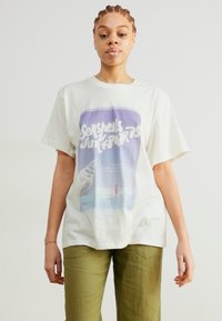 Lichtbeige katoenen t-shirt met een grafische print van een schelp en strandscène in paarse en blauwe tinten. Relaxte pasvorm, korte mouwen.