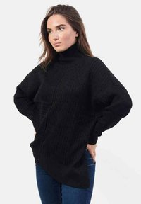 Maglione nero a collo alto lavorato a maglia con texture a coste, spalle scese, orlo asimmetrico e maniche larghe, abbinato a jeans in denim blu.