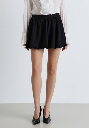 Frau trägt schwarze, aufgebauschte Shorts, weiße Bluse mit Rüschen vorne und schwarze spitze High Heels, steht vor einem einfarbigen Hintergrund.