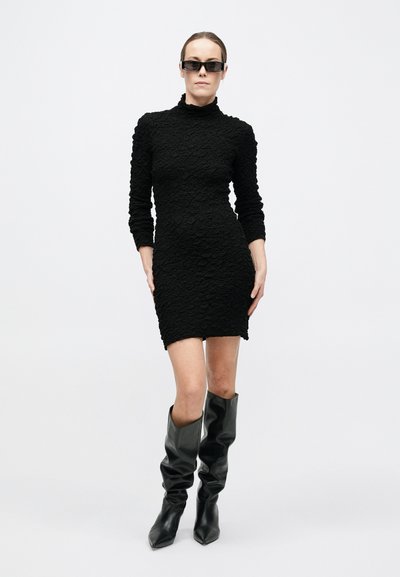 Naine, kes kannab tekstuuriga musta pika varrukaga minikleiti, musti lõdvaid põlvikukaitsmeid ja kandilisi musta päikeseprille valgel taustal.