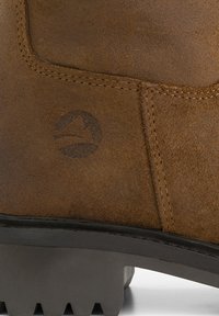 Gros plan du talon d’une botte en daim marron avec une semelle en caoutchouc noir et un logo montagne embossé sur le côté.