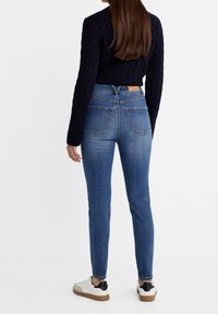 Jeans de mezclilla azul ajustados con cintura mid-rise, dos bolsillos traseros y una etiqueta de marca marrón en la cinturilla; combinados con un suéter negro de punto trenzado.