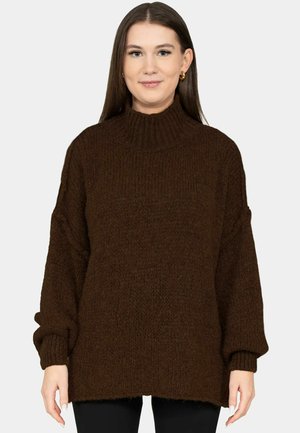 Frau mit langen braunen Haaren, die goldene Creolen und einen groben, dunkelbraunen Rollkragenpullover trägt, steht vor einem schlichten hellen Hintergrund.