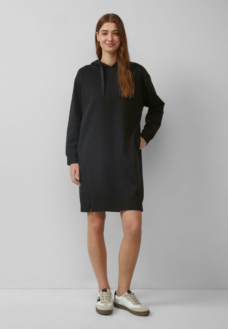 Schwarzes Hoodie-Kleid mit langen Ärmeln, Rippbündchen und seitlichen Schlitzen. Weicher Stoff mit entspannter Passform, kombiniert mit lässigen Turnschuhen.