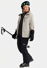 Lichtgrijze ski-jas over een zwarte geïsoleerde laag, gecombineerd met zwarte broek en handschoenen. Accessoires zijn onder andere een zwarte helm en skistokken.