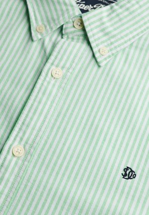 Camisa abotonada con rayas verticales verdes y blancas, botones beige y pequeño logo bordado en negro cerca de la parte inferior derecha.