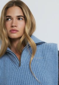 Chaleco de punto azul claro con un alto cuello y media cremallera. Presenta un patrón texturizado y un ajuste relajado. Cabello estilizado en ondas sueltas.