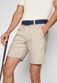 Béžové bavlnené šortky s bočnými vreckami, so spleteným námorníckym pásom a plochým predným dizajnom. V jednej ruke drží čierny golfový palica.