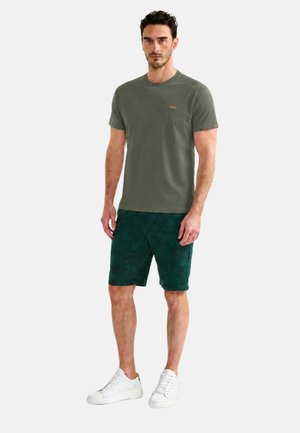 Uomo in piedi che indossa una maglietta verde oliva, pantaloncini verde scuro a fantasia e sneaker bianche su sfondo bianco semplice.