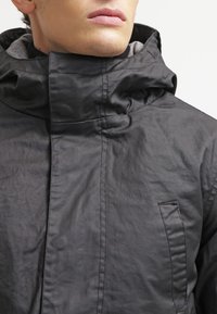 Homme portant une veste à capuche noire avec un col haut, gros plan montrant la texture du tissu et les détails de couture.