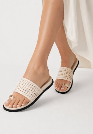 Bare fødder iført beige vævede sandalslipper med tå-loop og gulddetalje, kombineret med en flydende hvid kjole mod en ensfarvet baggrund.