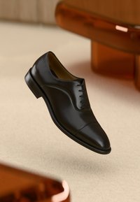 Zapato de vestir de cuero negro con un acabado suave, punta redonda y cordones clásicos. Se encuentra sobre una superficie ligera y texturada.
