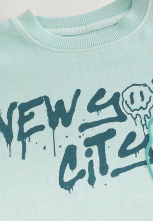 Light blue cotton t-shirt featuring dark blue graffiti-style text “NEW YORK CITY” with drips and round smiley graphic accents.
