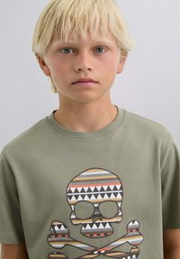 Camiseta de algodón verde oliva con un diseño de calavera y huesos cruzados en tonos marrón, negro, blanco y rojo. Mangas cortas y cuello redondo.