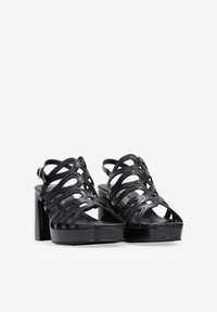 Black high-heeled sandals with open toe, intricate cut-out design, and platform sole. Made from glossy material with a chunky heel.