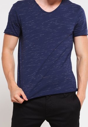 Uomo che indossa una t-shirt blu navy a maniche corte con sottili strisce bianche, tira leggermente l'orlo, vestito con pantaloni neri.