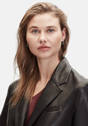Zwarte leren blazer met scherpe revers, textuurafwerking, gecombineerd met een bordeaux top en zilveren oorbellen met een hoop. Haar is middellang en golvend.