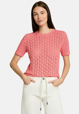 Woman with straight brown hair wearing a coral pink textured short-sleeve top and white pants with a drawstring, standing with hands in pockets.