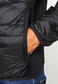 Svart puffjacka med en slät yttermaterial, med kviltade sektioner, sidofickor med dragkedja och ett mjukt kontrasterande innerfoder.