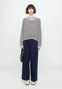 Camicia a righe con maniche lunghe in crema e blu navy, abbinata a pantaloni larghi blu navy e sneakers bianche. Tiene una borsa nera texture.