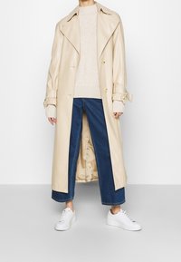Trench-coat en cuir beige avec col surdimensionné, boutons croisés et manches larges, associé à un pull léger et un jean bleu raccourci.