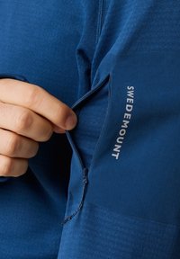 Giacca blu con zip in un tessuto testurizzato. Presenta una tasca laterale e un'etichetta "SWEDEMOUNT" bianca sulla parte superiore del braccio.