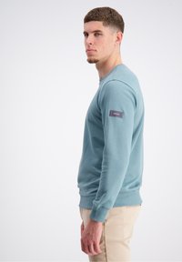 Ljusblå sweatshirt i mjukt material, rund halsringning, ribbad ärmslut och nederkant, med en tyglabel på vänster ärm. Stylad med beige byxor.