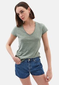 T-shirt verde a maniche corte con scollatura a V realizzato in tessuto leggero, abbinato a pantaloni corti in denim blu con orlo arrotondato e tasche visibili.